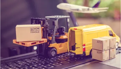 A miniature forklift carries a box toward a yellow delivery van, with stacks of boxes nearby and an airplane flying overhead, all set on a laptop keyboard, representing logistics or ecommerce.